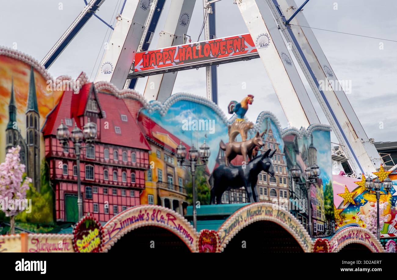 Bremen, Deutschland. 31. Oktober 2025. Die Worte „Happy Halloween“ laufen über eine LED-Videowände am Riesenrad auf der Bürgerweide, während die Bremer Stadtmusiker an einem Stand im Vordergrund zu sehen sind. Der Bremer Freimarkt ist eines der ältesten Volksfeste in Deutschland und eines der größten in Norddeutschland. Quelle: Hauke-Christian Dittrich/dpa/Alamy Live News Stockfoto