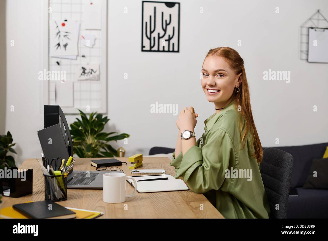 Die junge Frau lächelt glücklich in ihrem Zuhause, während sie an ihrem Schreibtisch kreativ arbeitet. Stockfoto