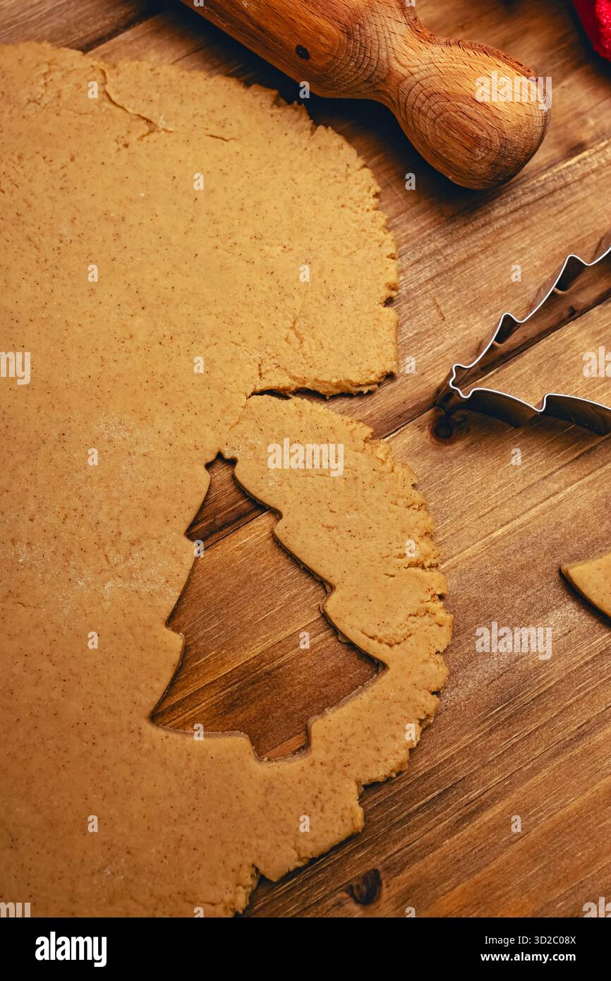Auf einem Holztisch wird ausgerollter Keksenteig mit Weihnachtsbaumform dargestellt, der mit einem Keksschneider aus Metall ausgeschnitten wurde. Stockfoto