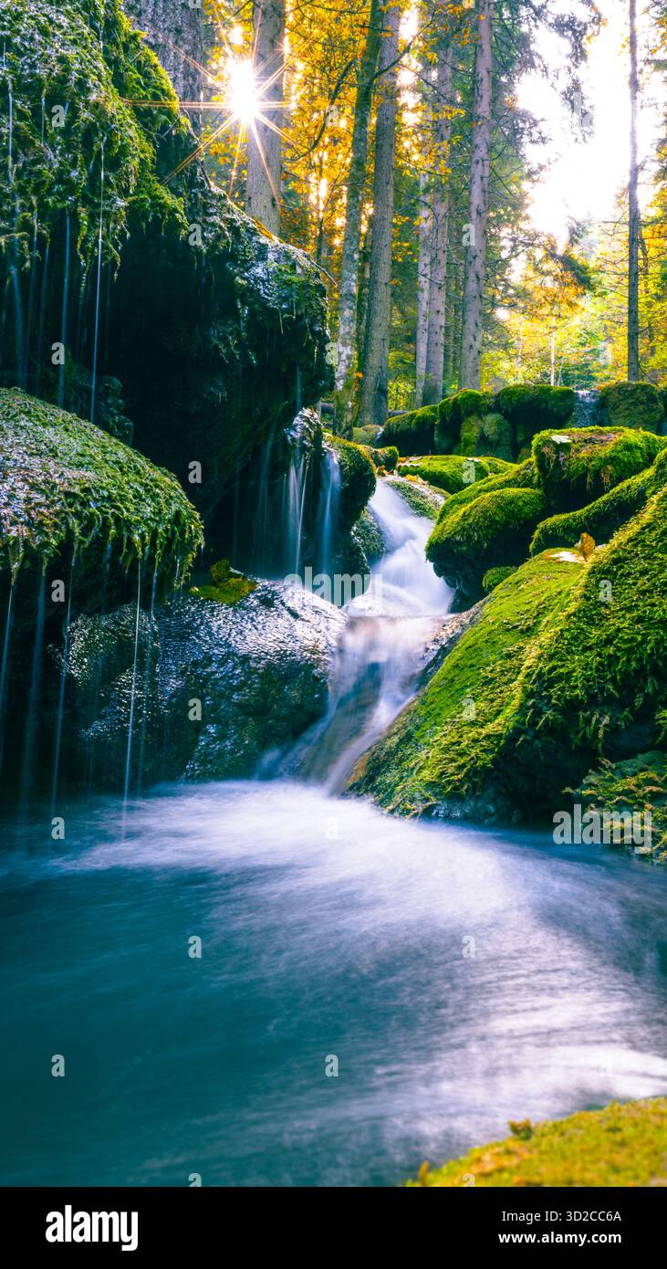 Moosige Felsen und Wasserfall, beleuchtet von Sonnenlicht, das durch dichten Herbstwald strömt Stockfoto