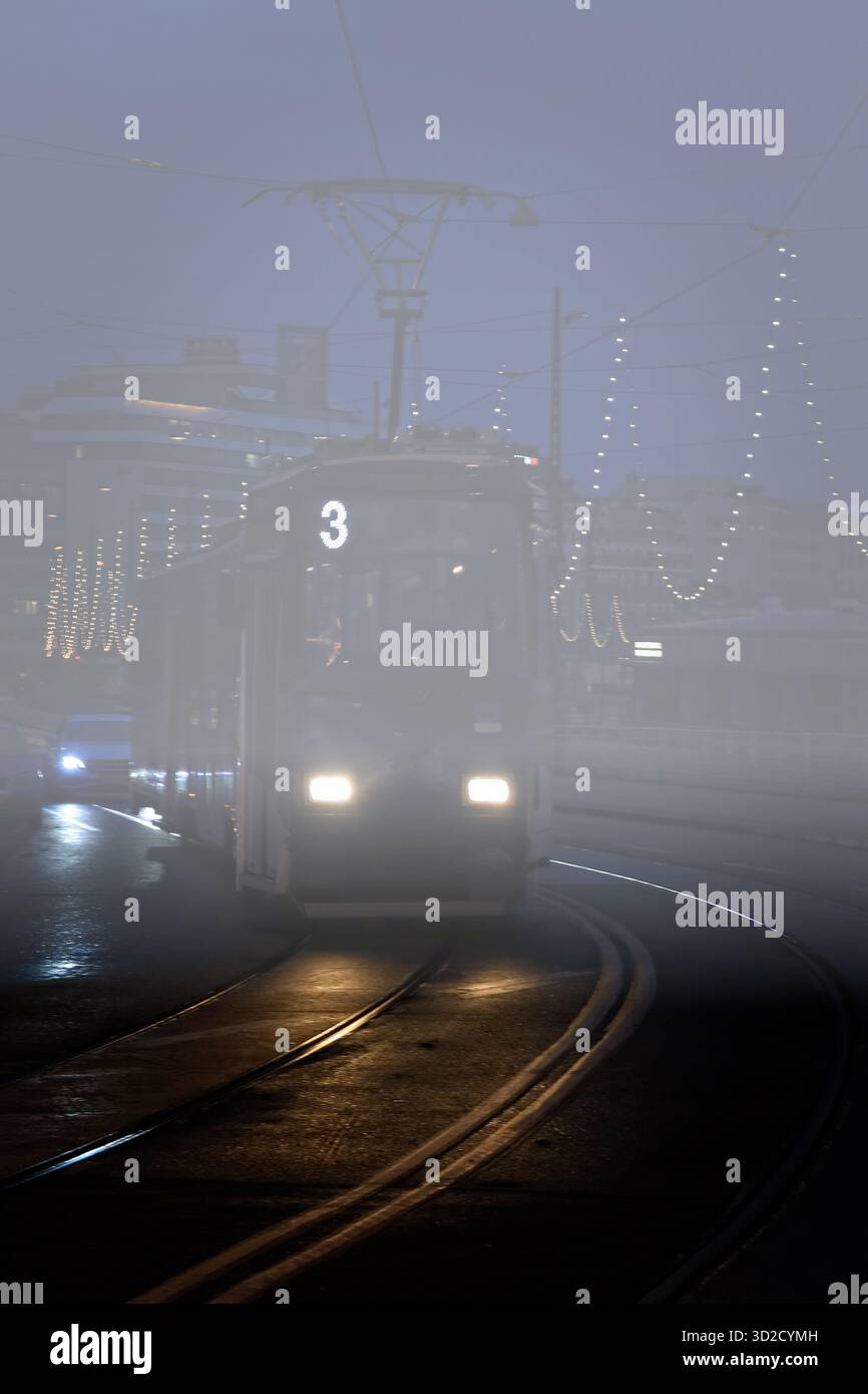 Die Straßenbahn kommt an einem dunklen, nebeligen Novembermorgen zur Straßenbahnhaltestelle. Helsinki, Finnland. Stockfoto