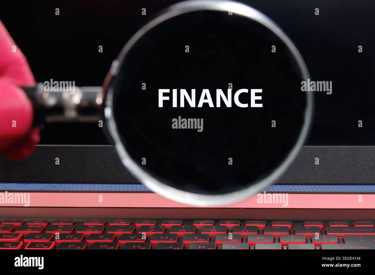 Eine Lupe wird an einem Laptop-Bildschirm gehalten, wodurch das Wort Finanzen hervorgehoben wird. Die Tastatur leuchtet rot auf und symbolisiert einen fokussierten Blick auf die Busine Stockfoto