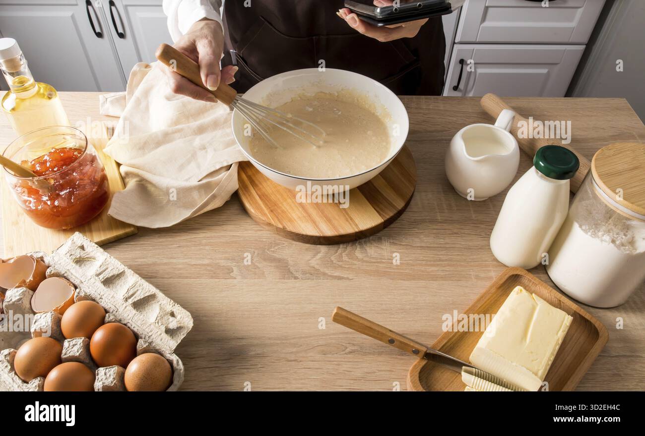 Die Hand einer Frau mit einem Schneebesen schlägt den Teig in einer Schüssel. Die andere Hand hält das Telefon mit dem Rezept für das weitere Kochen Stockfoto
