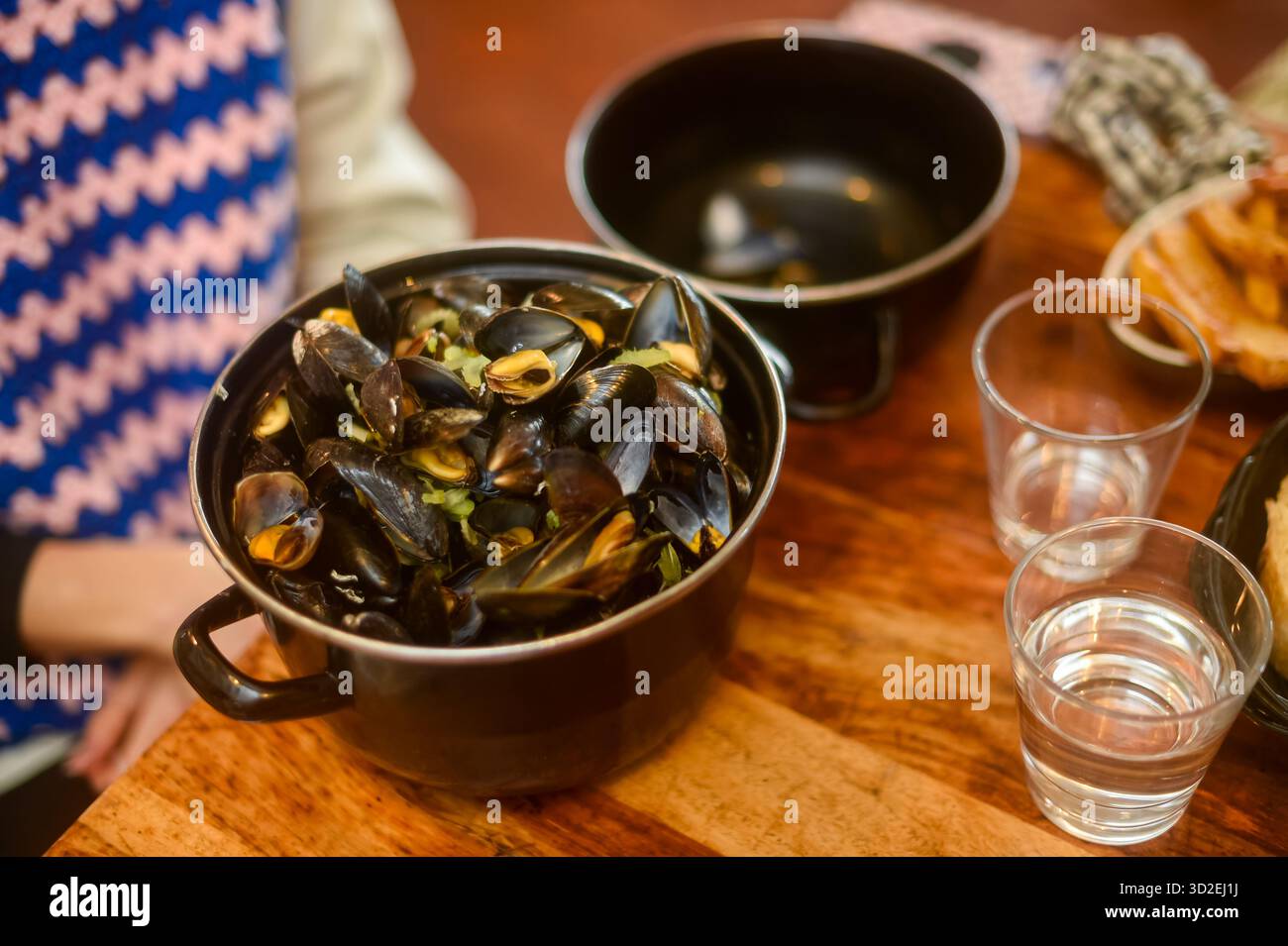 Ein Topf mit heißen, frisch gekochten Muscheln in Soße steht auf einem Holztisch, neben zwei Gläsern Wasser oder Wein. Bietet traditionelle französische Küche und ein warmes, gemeinsames kulinarisches Erlebnis. Horizontal. Stockfoto