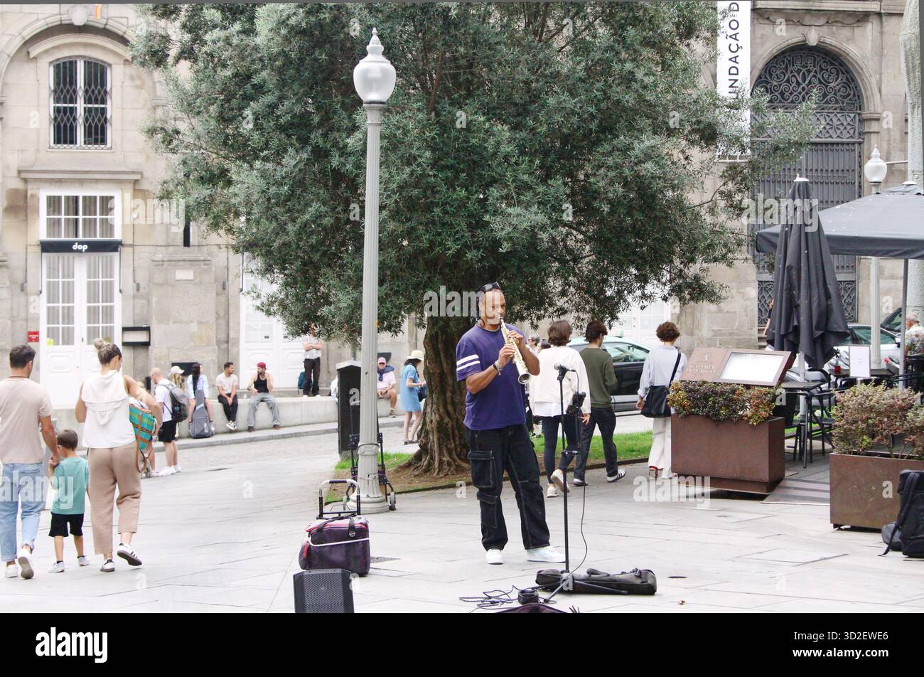 Porto, Portugal - 10. Juni 2025: Musiker spielt Sopransaxophon auf der belebten plaza, umgeben von Bäumen und Menschen, die die lebhafte Straßenszene genießen Stockfoto