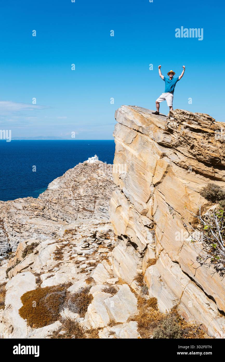 Ein Mann steht in einer Siegespose auf einem felsigen Gipfel vor dem Leuchtturm im Paros Park Nature Reserve, Griechenland. Stockfoto