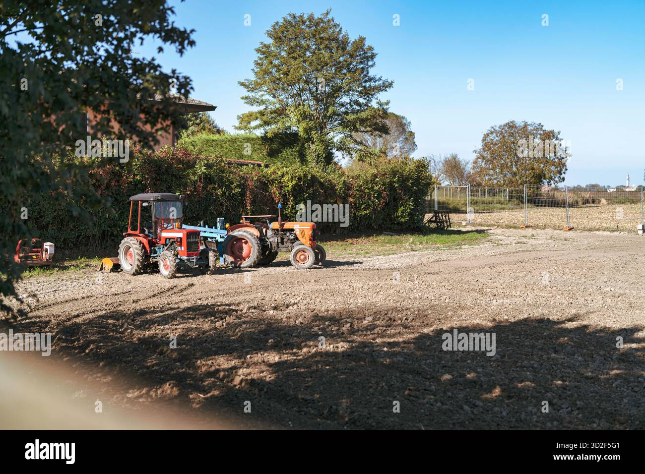 Zwei klassische landwirtschaftliche Traktoren mit landwirtschaftlichen Arbeitsgeräten stehen auf einem sonnendurchfluteten Feld unter klarem blauen Himmel und demonstrieren die Arbeit auf dem Bauernhof und Stockfoto