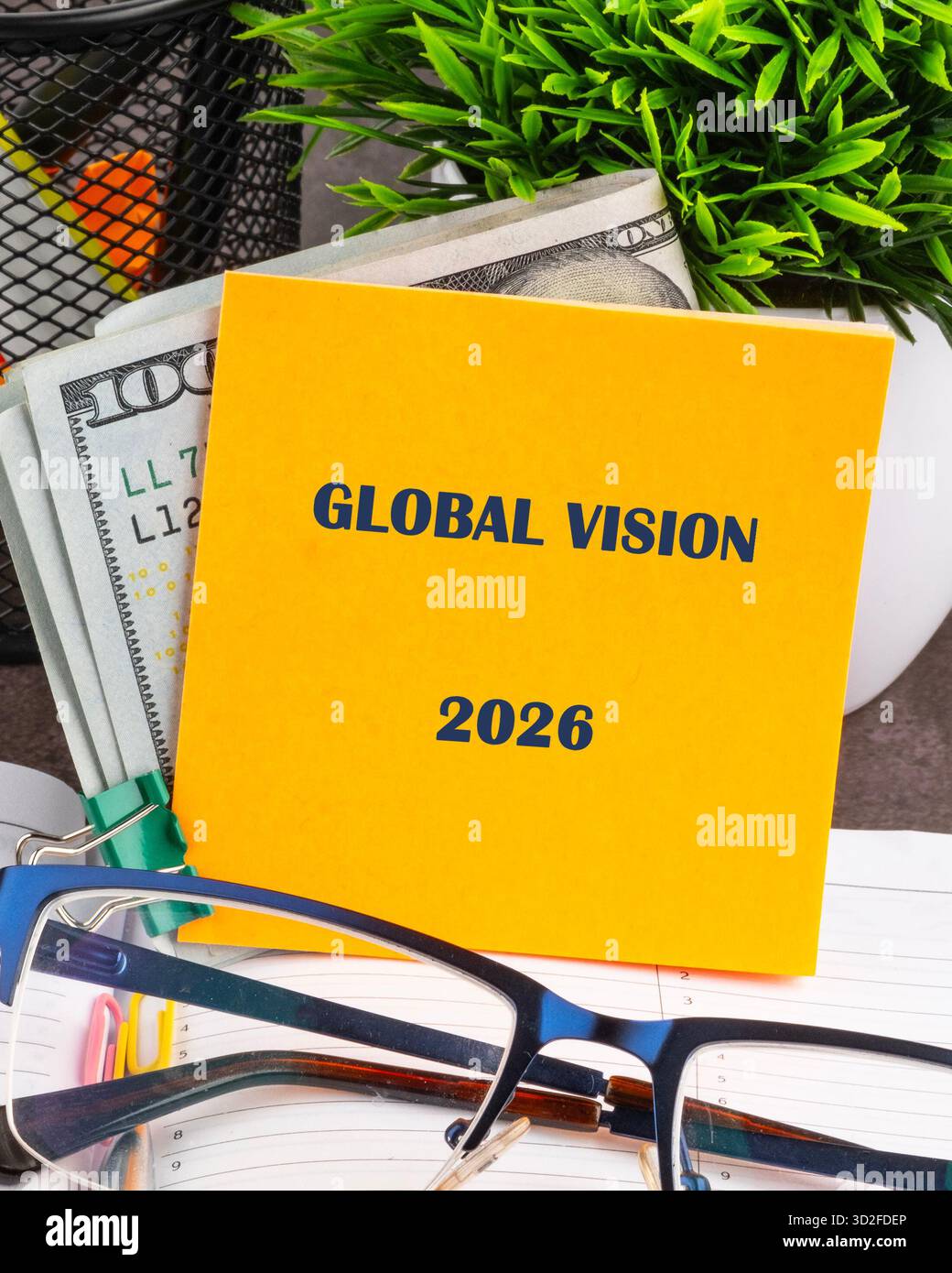 Gelbes Notizbuch mit Global Vision 2026 ist neben US-Währung, Brillen, einer kleinen grünen Pflanze und einem Organisator auf der Oberseite eines Planers. Futu Stockfoto