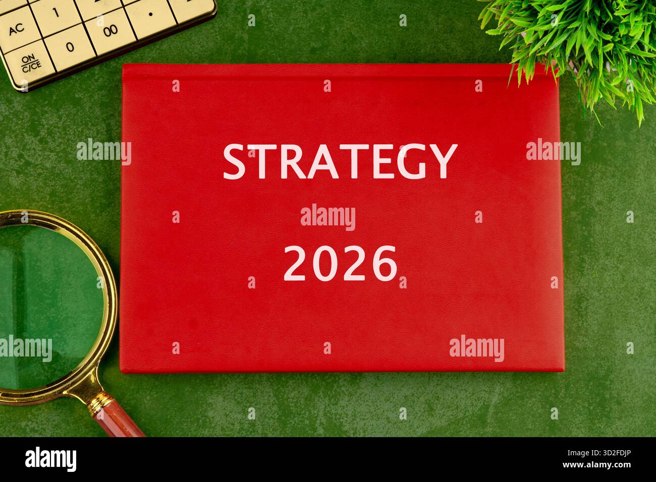 Ein rotes Buch mit dem Titel Strategie 2026 sitzt auf einer strukturierten grünen Oberfläche. Es gibt einen Taschenrechner, eine Lupe und eine grüne Pflanze im Blick. Stockfoto