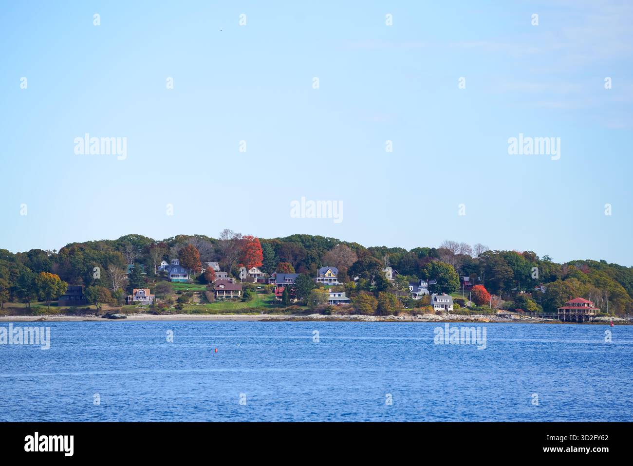 Ein landschaftlich reizvolles Küstenviertel mit Blick über das ruhige blaue Wasser, farbenfrohe Herbstbäume und Häuser am Wasser unter hellem, offenem Himmel. Stockfoto