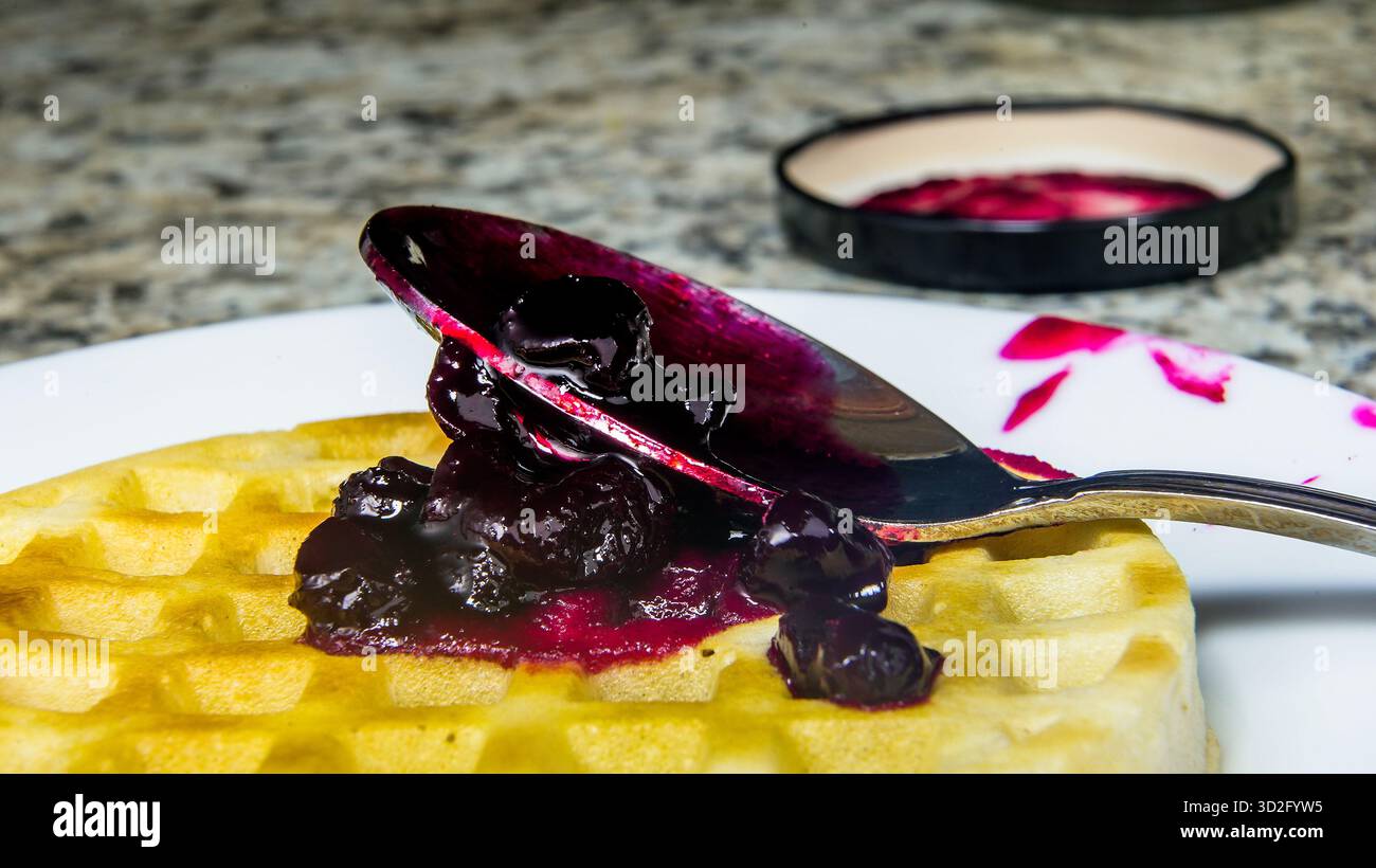 Eine flauschige Waffel wird auf einem weißen Teller mit großzügigem wildem Huckleberry-Aufstrich platziert. Ein glänzender Löffel liegt auf der Waffel, bereit für einen leckeren Happen, in einem warmen K Stockfoto