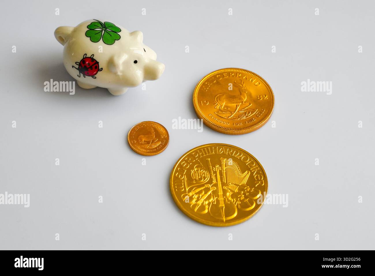 Piggy Bank mit Lucky Shamrock und Marienkäfer, umgeben von glänzenden Goldmünzen aus der „Krugerrand“- und „Philharmoniker“-Serie, auf Einem Plain Gray Backgrou Stockfoto