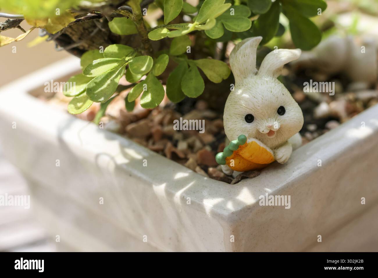 Niedliche weiße Miniatur-Kaninchen-Figur mit einer orangen Karotte sitzt in einem kleinen Gartentopf. Diese charmante Dekoration zwischen grünen Pflanzen schafft skurrile, Stockfoto