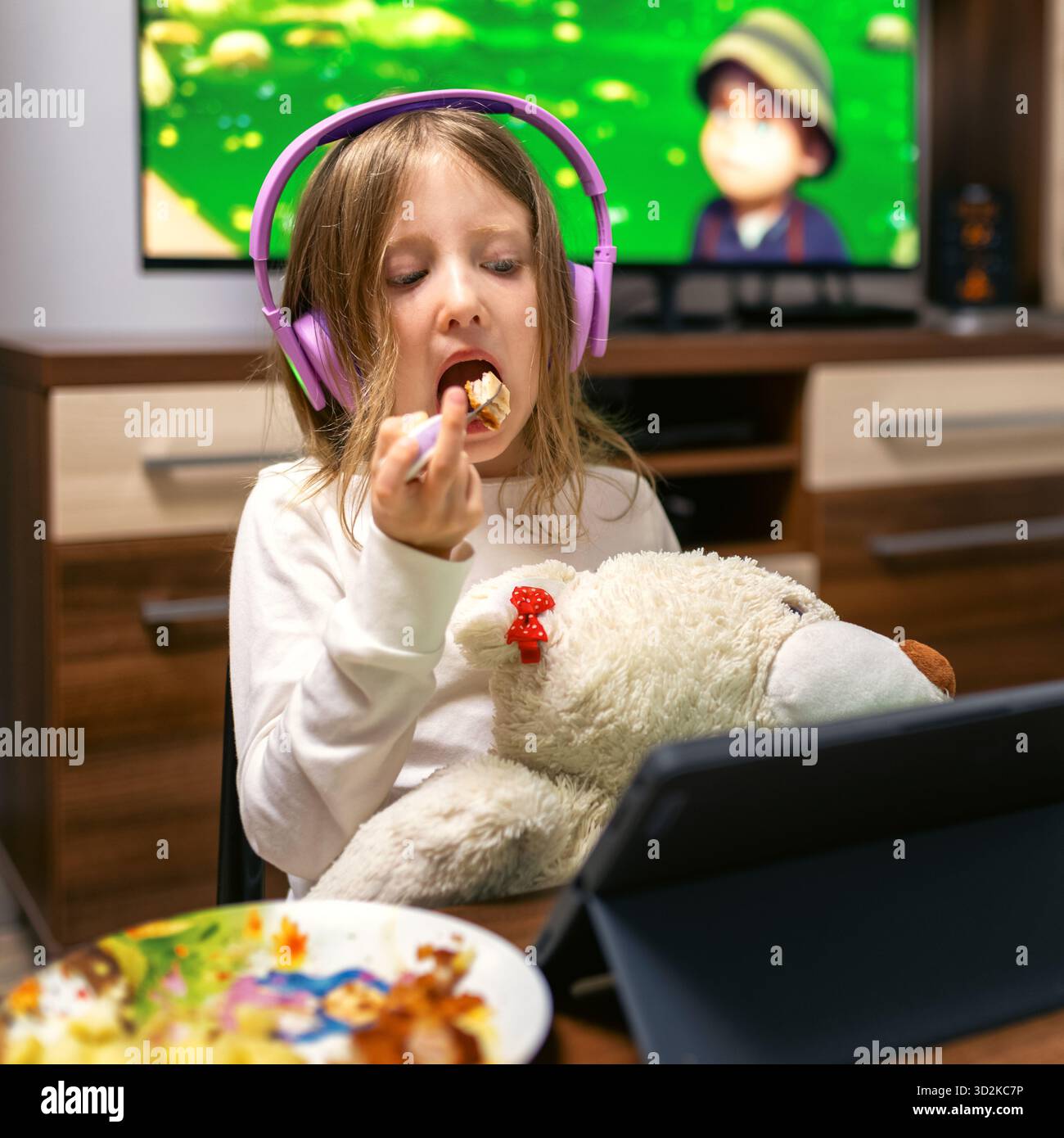 Kind mit violetten Kopfhörern beim Mittagessen, während es ein Tablet ansieht. Konzept zur Darstellung digitaler Ablenkung, Bildschirmabhängigkeit und ungesunder Gewohnheiten in c Stockfoto