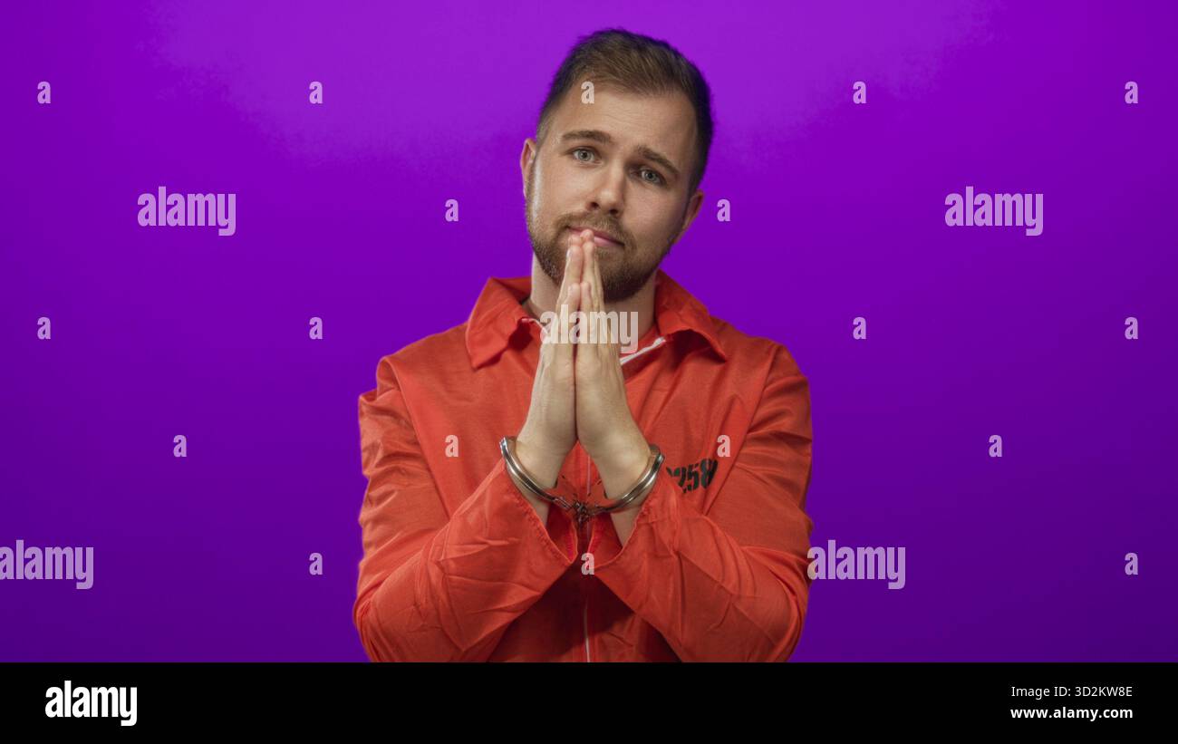 Mann, junger kaukasischer Gefangener in orangefarbenem Jumpsuit mit Handschellen, in Gebetsgeste an einer violetten Atelierwand gehalten; Reue. Stockfoto