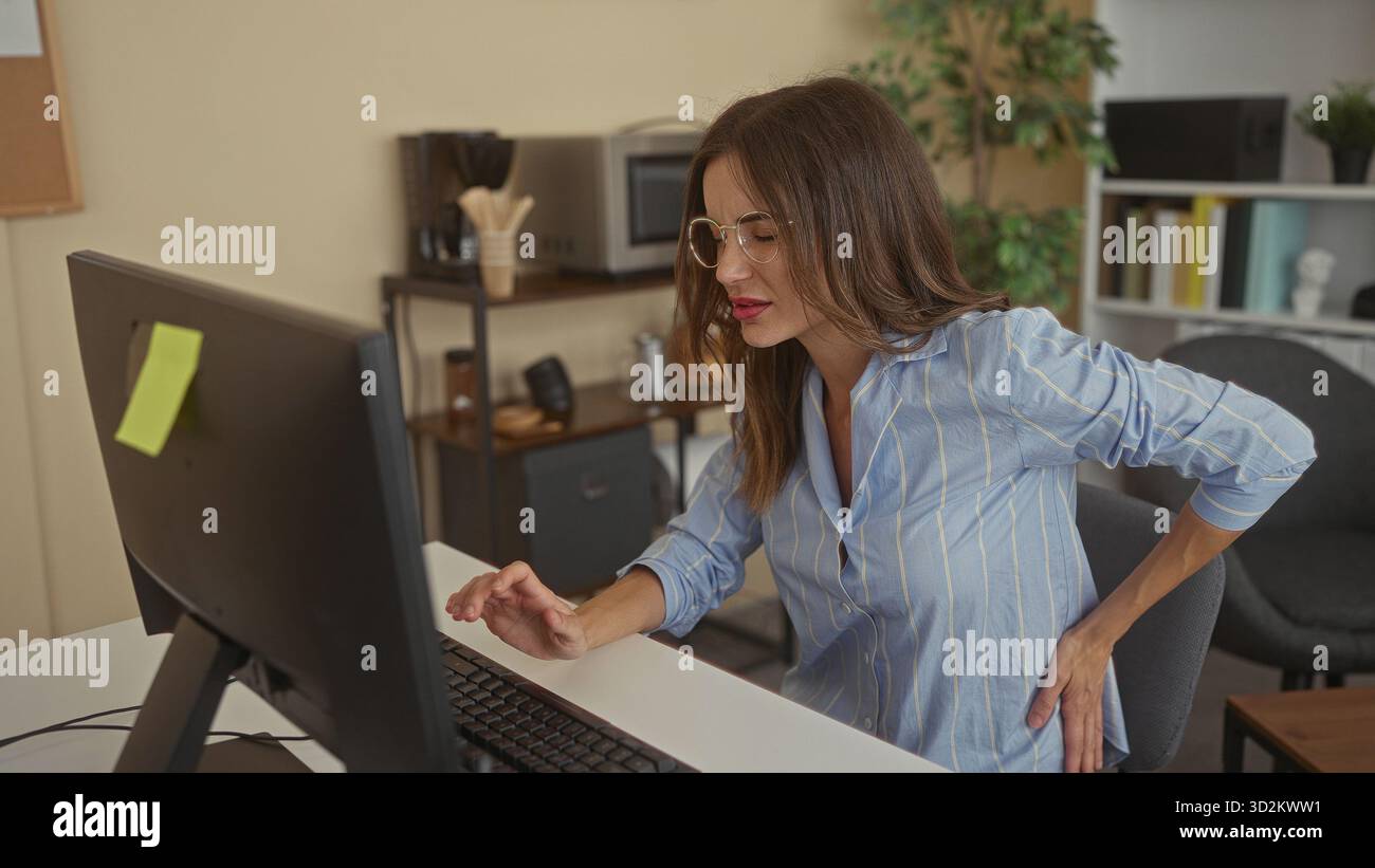 Frau mit Brille hält den unteren Rücken, während sie auf der Desktop-Tastatur am Schreibtisch des Büros schreibt; Schmerzen. Stockfoto