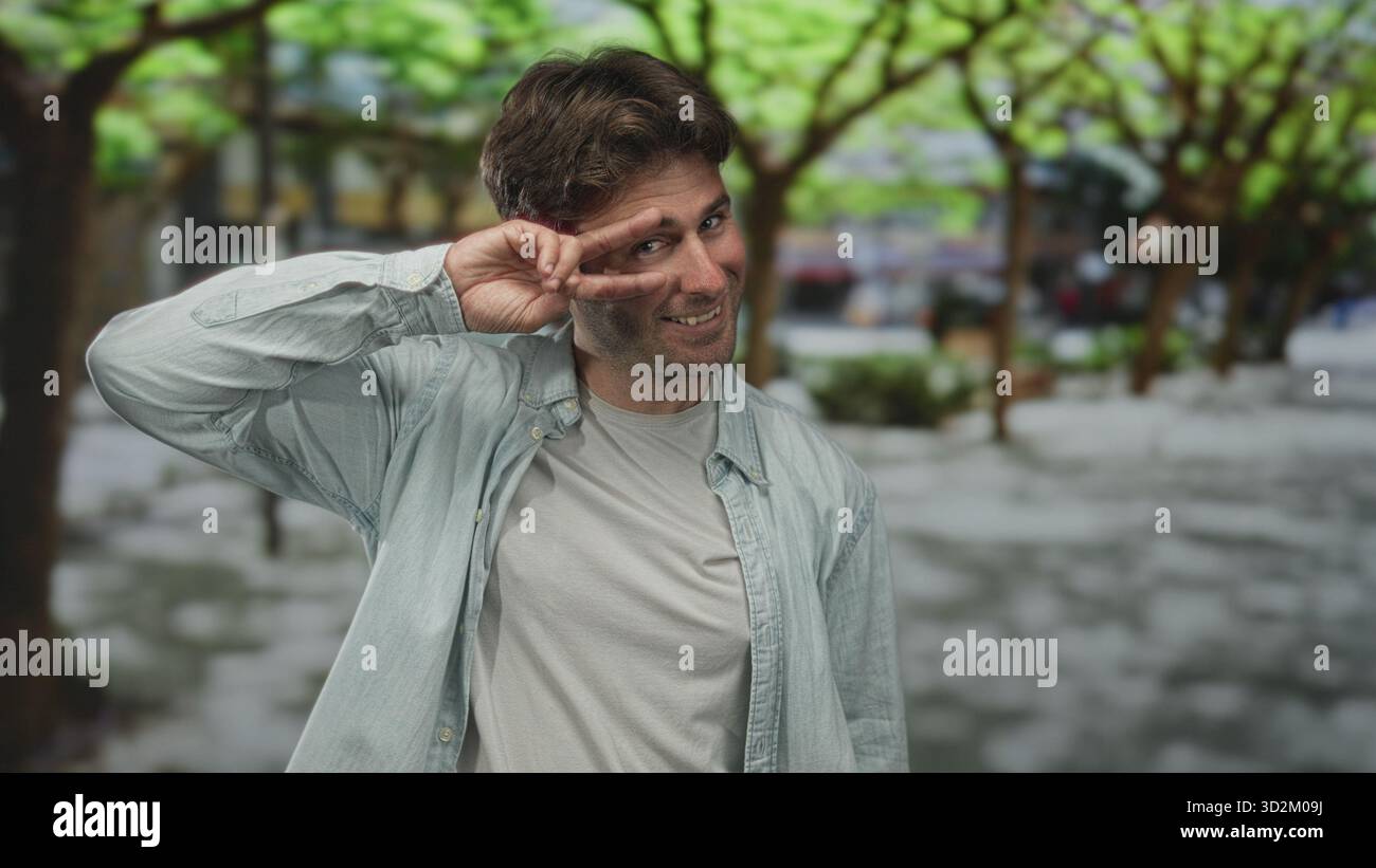 Der Mann hält zwei Finger in einem Friedensschild auf die von Bäumen gesäumte Straße, lächelt in einem hellen Hemd über weißem T-Stück; verspielte Freizeit. Stockfoto
