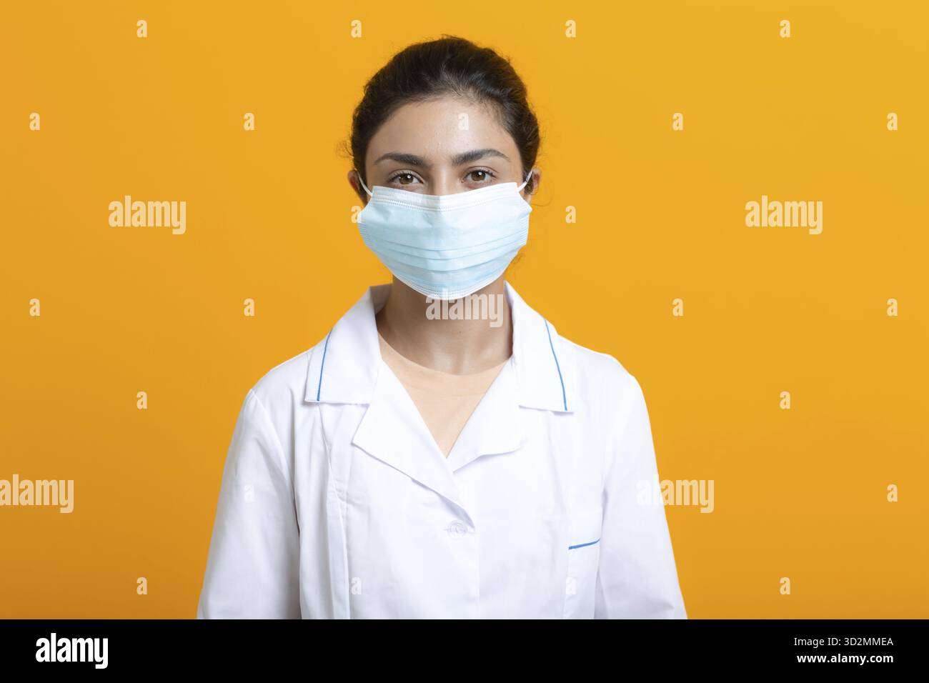 Porträt der indischen Ärztin, die weißes medizinisches Kleid und Gesichtsmaske trägt, isoliert auf gelbem Hintergrund Stockfoto