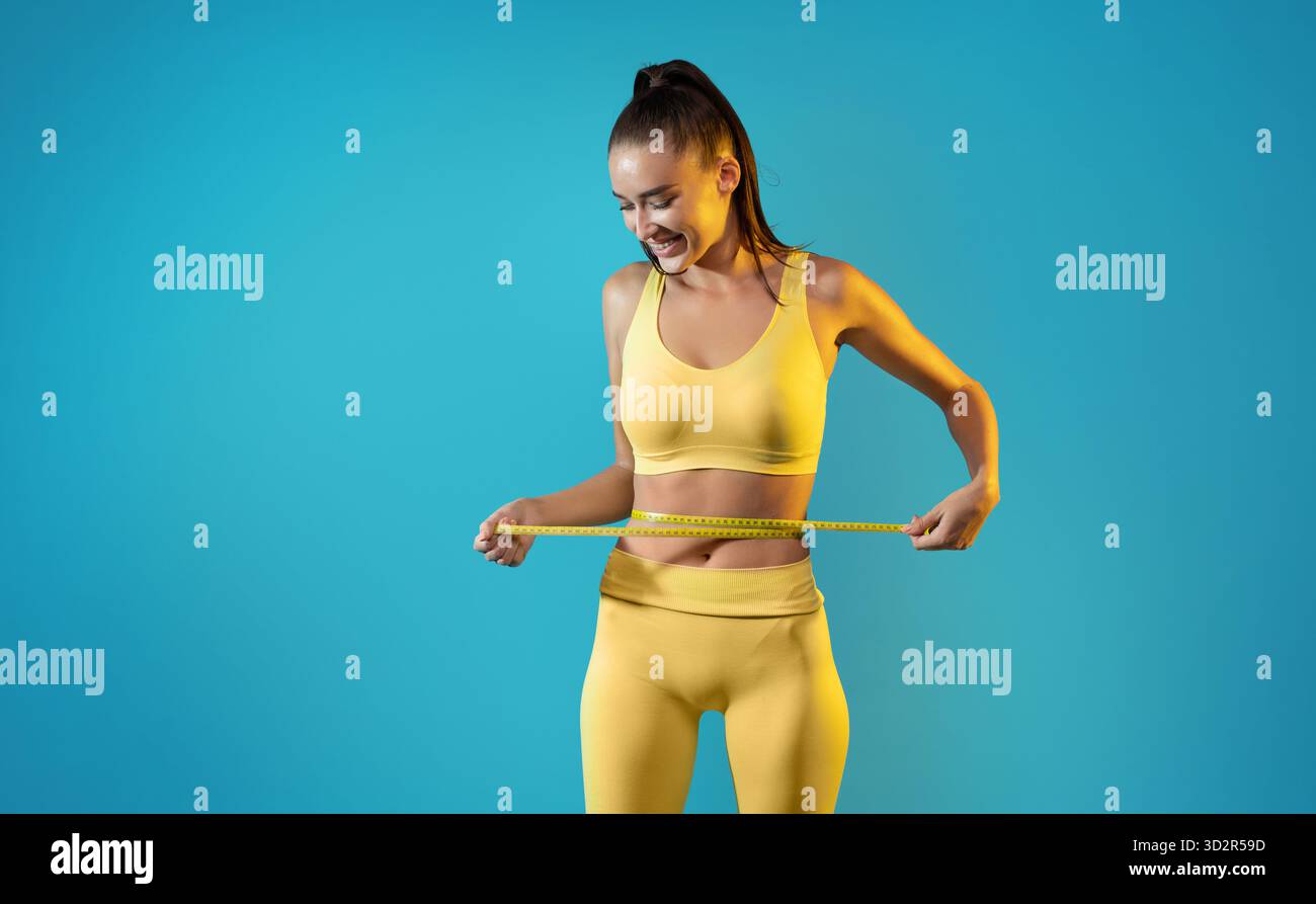 Sportliche Woman Measuring Taille After Weight Loss Success in Bright Blue Studio Stockfoto