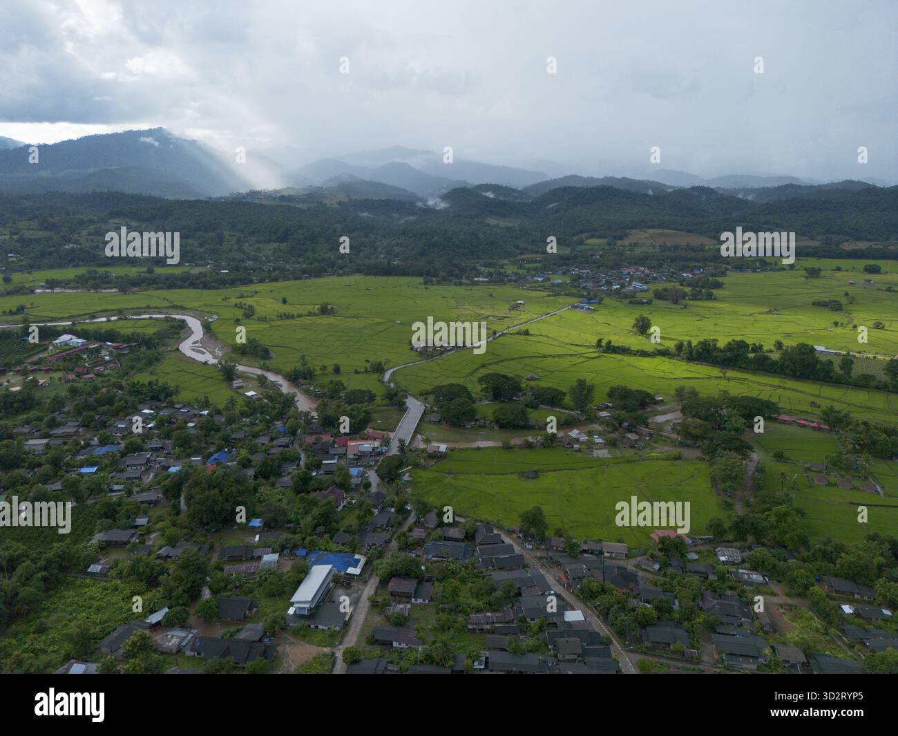 Die weitläufige Luftlandschaft zeigt ein friedliches ländliches Dorf, eingebettet zwischen grünen Feldern, mit einem sich windenden Fluss, der in ferne Berge fließt und bedeckt ist Stockfoto
