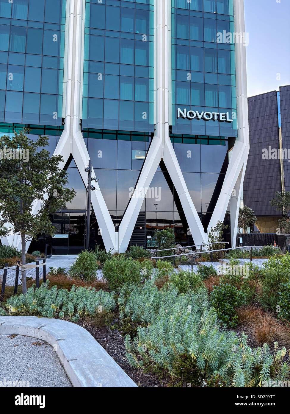 Novotel Auckland Airport Hotel. Modernes, glasverkleidetes Gebäude mit markanten weißen geometrischen Stützen und zeitgenössischer Landschaftsgestaltung am Eingang. Stockfoto