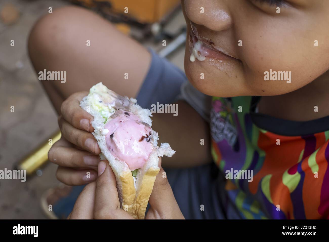 Nahaufnahme eines unordentlichen asiatischen Kindes, das glücklich süßes rosafarbenes Eisbrot-Sandwich isst. Dieses offene Foto zeigt, wie das Kind das Dessert genießt, mit einer Ameise auf dem Essen Stockfoto