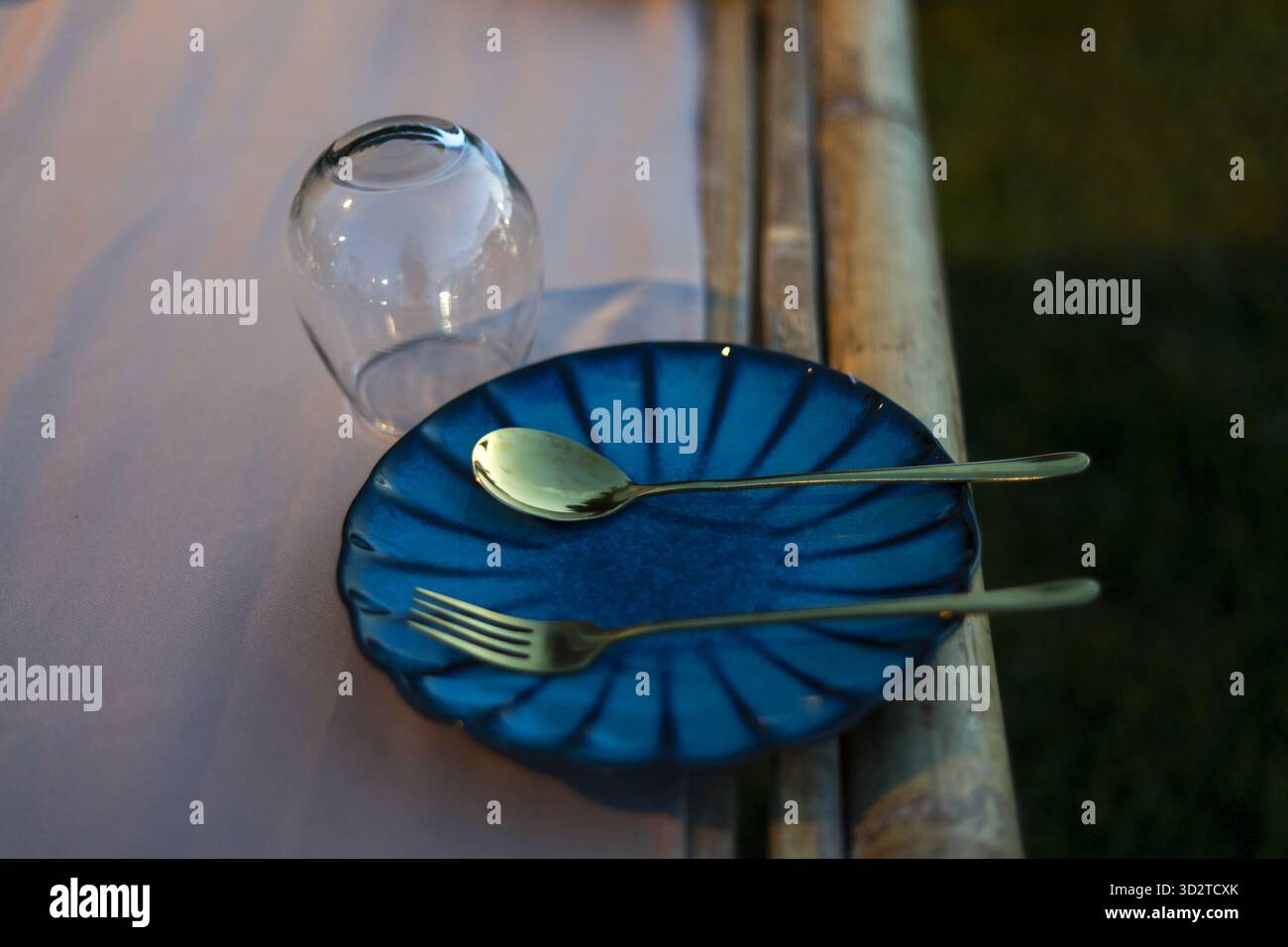 Romantischer Esstisch mit eleganter blauer Keramikplatte und goldenem Besteck. Löffel und Gabel bereit für eine intime Mahlzeit auf rustikaler Bambusoberfläche Stockfoto
