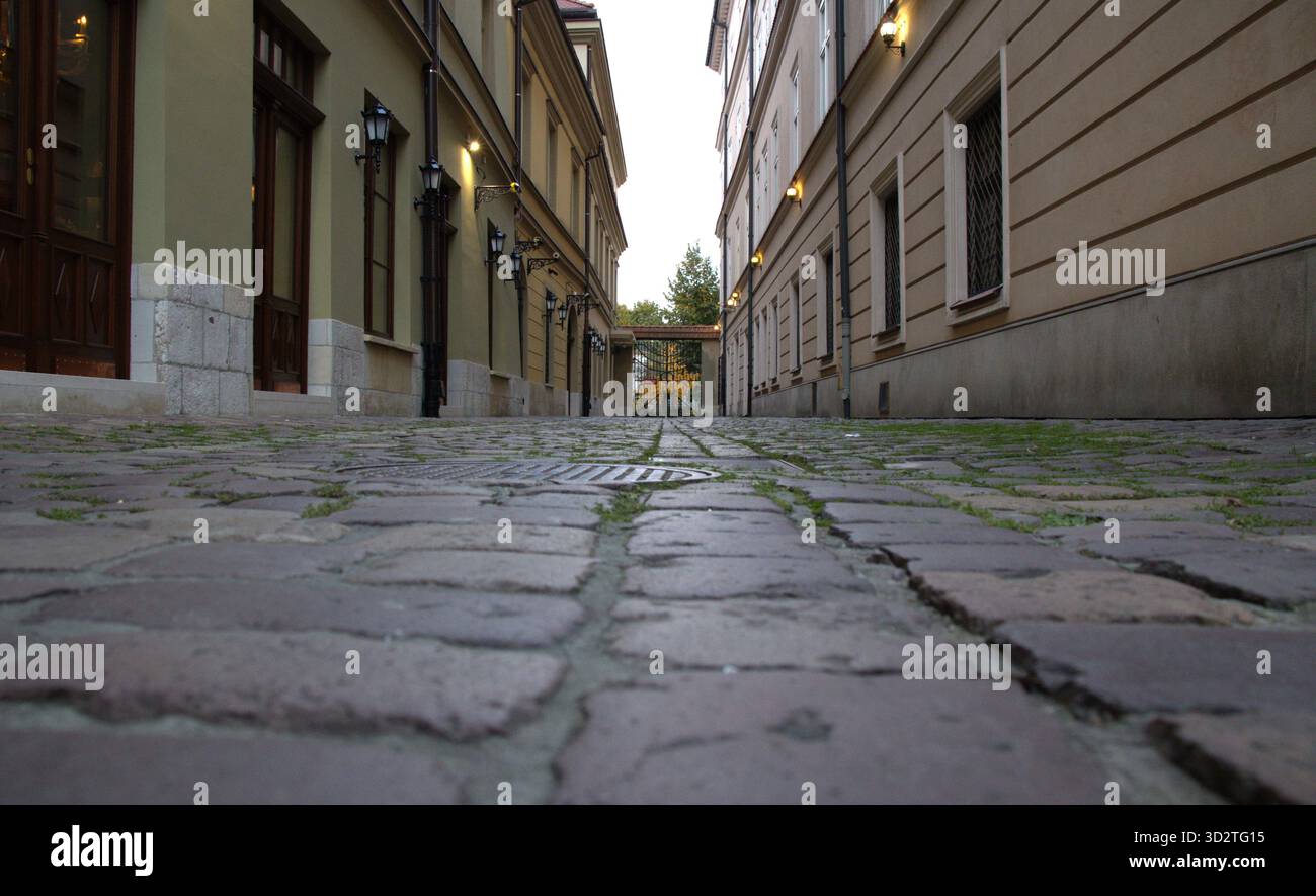 Ein ebenerdiger Blick auf eine enge, leere Gasse, die mit abgenutzten Kopfsteinpflastern gepflastert ist. Historische Gebäude flankieren beide Seiten mit Holztüren. Krakau, Polen Stockfoto