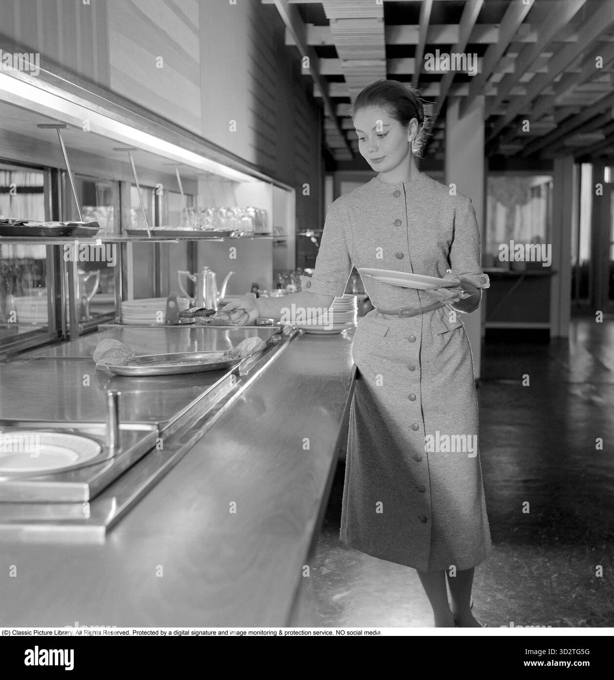 Sie kleidete sich schön 1956. Das junge Model hilft sich beim Essen im Restaurant. Sie trägt die typische Kleidung der Periode, die Hemdkleid oder Hemdkleid genannt wird. November 1956 Stockfoto