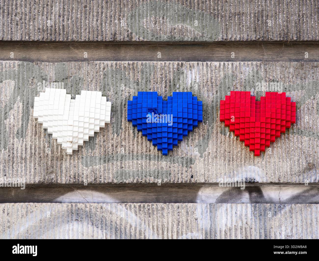 Drei gepixelte Herzen in weiß, blau und rot auf einer strukturierten grauen Wand angeordnet Stockfoto
