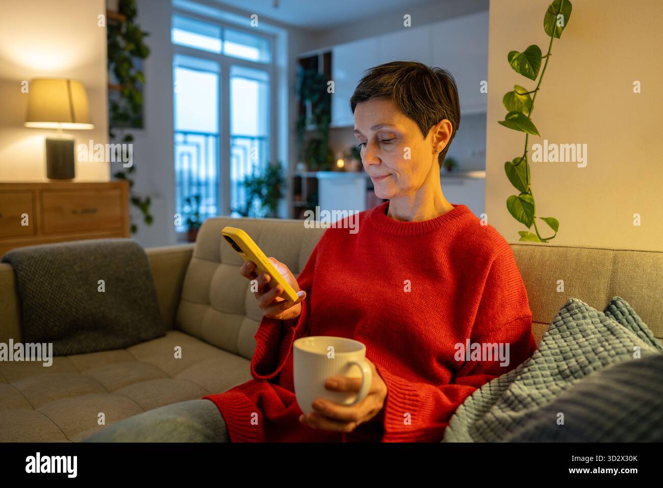 Seniorin ruht auf der Couch im Wohnzimmer, hält Kaffeetasse und blättert Medien auf dem Smartphone. Stockfoto