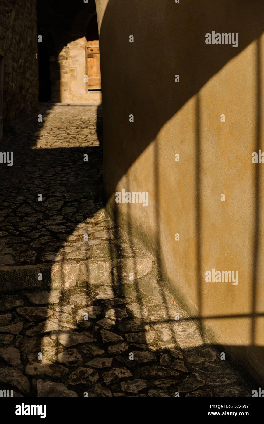 Goldenes Sonnenlicht wirft lange künstlerische Schatten auf eine enge Kopfsteinpflasterallee. Minimalistisches Spiel von Licht, Schatten und Textur in einer alten europäischen Stadt Stockfoto