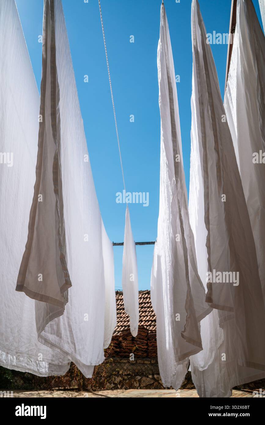 Waschtag unter hellblauem Himmel. Klare weiße Blätter wehen sanft in der Sonne und schaffen dramatische Linien gegen rustikales Terrakottadach und Steinmauer Stockfoto