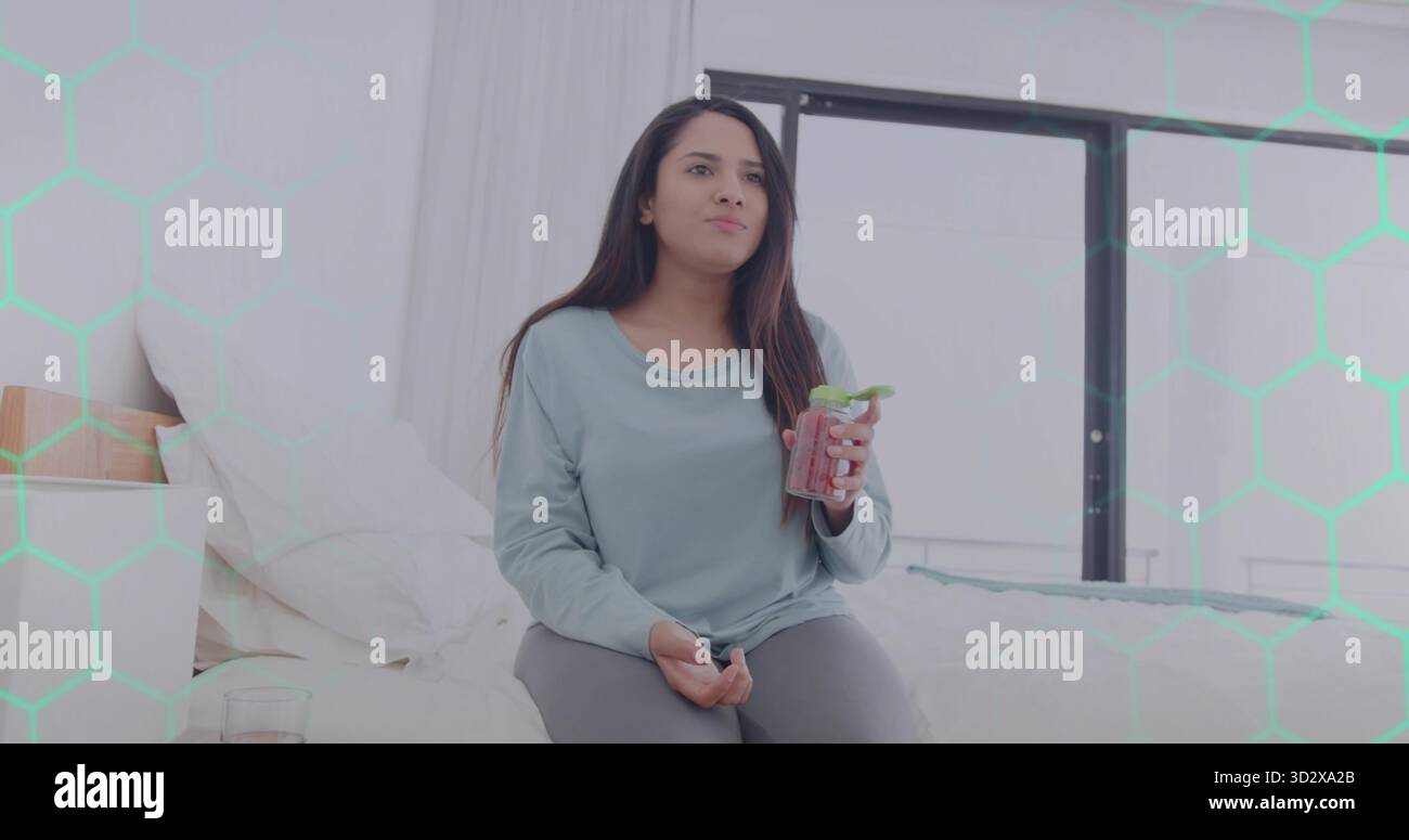 Sitzende indische Frau mit Mint-Smoothie-Dose in modernem Schlafzimmer mit Nachttisch und Fenster Stockfoto