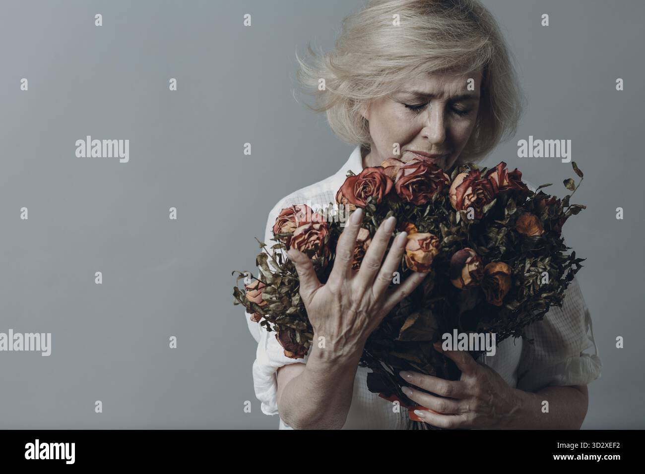 Traurige ältere Frau, die einen Strauß verwelkter Rosenblüten berührt. Grauhaarige reife Dame in Depression. Konzept des alten Alterns. Studioaufnahmen isoliert auf GRA Stockfoto