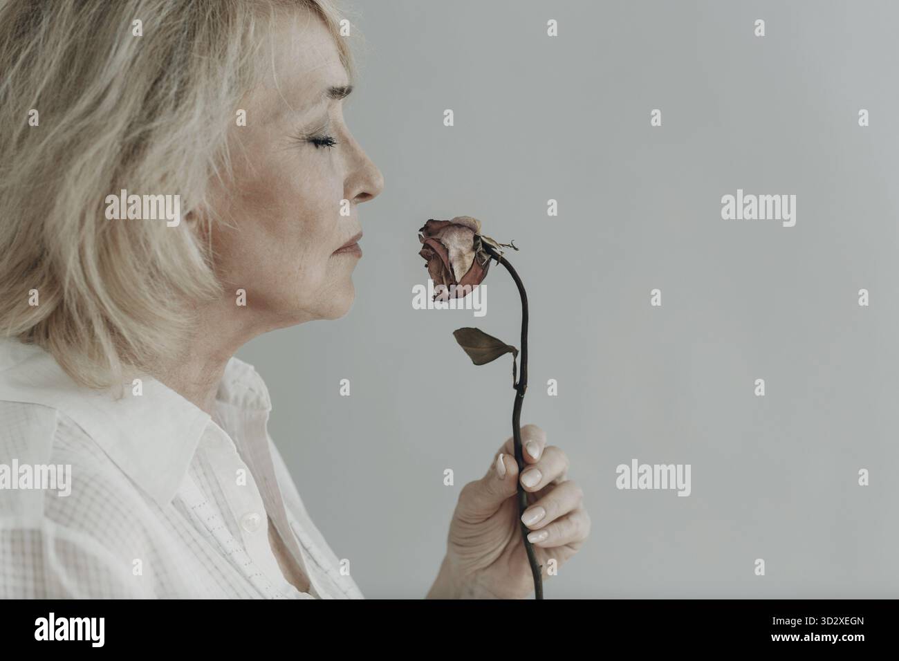 Traurige ältere ältere Frau in weißem Hemd hält eine trockene graue alte tote Rosenblume. Alter Konzept Stockfoto