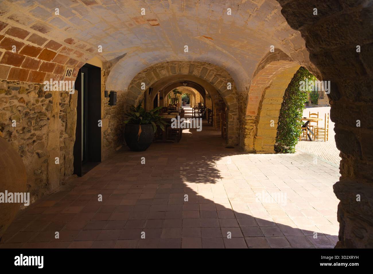 Steinbögen und rustikale Tische in einer schattigen mittelalterlichen Arkade in Monells, Katalonien, umgeben von warmem Sonnenlicht und historischem Charme. Stockfoto