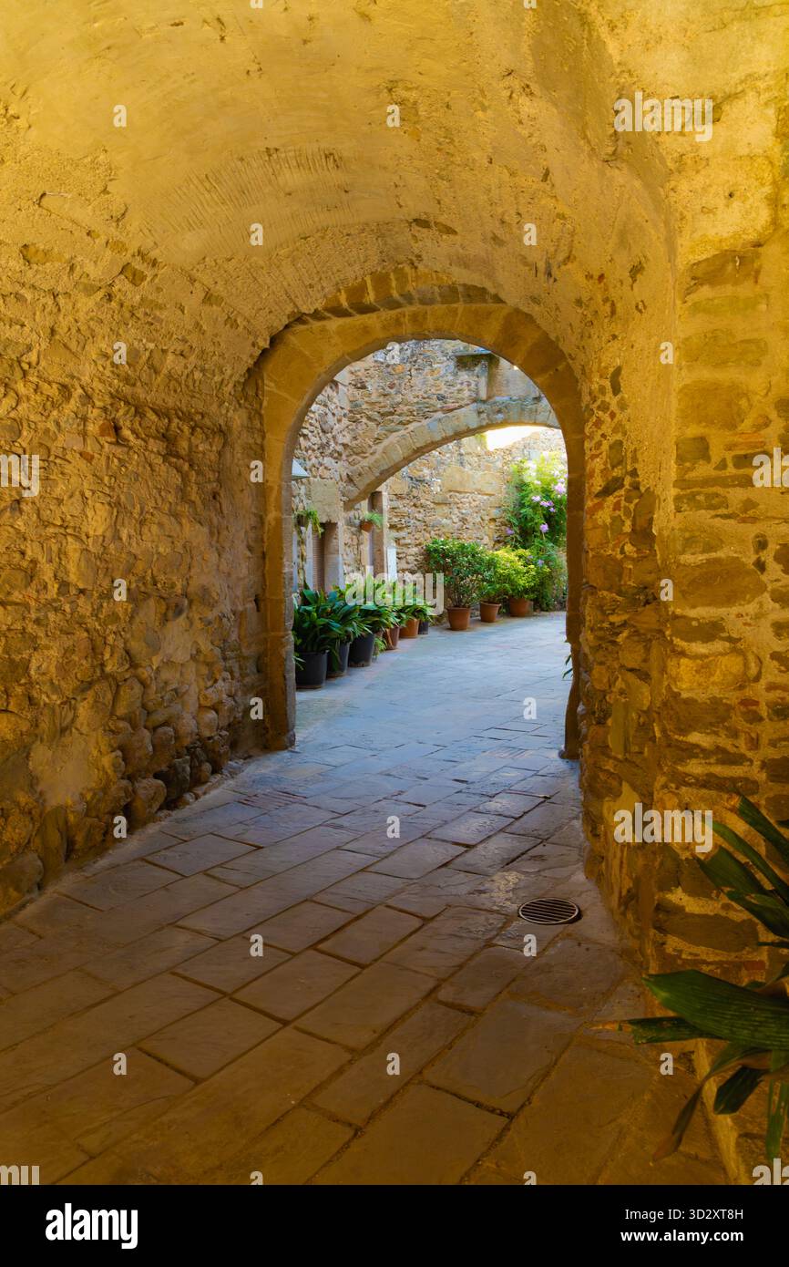 Steingasse mit Bögen in Monells, Katalonien, gesäumt von Topfpflanzen und in warmes Licht und historische Atmosphäre getaucht. Stockfoto