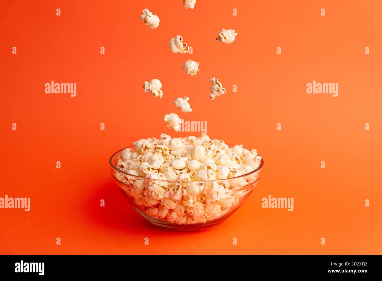 Popcorn fliegt über einer Glasschale auf orangem Hintergrund und symbolisiert Freude, Bewegung und filmische Momente in einer verspielten, kreativen Studiokomposition Stockfoto