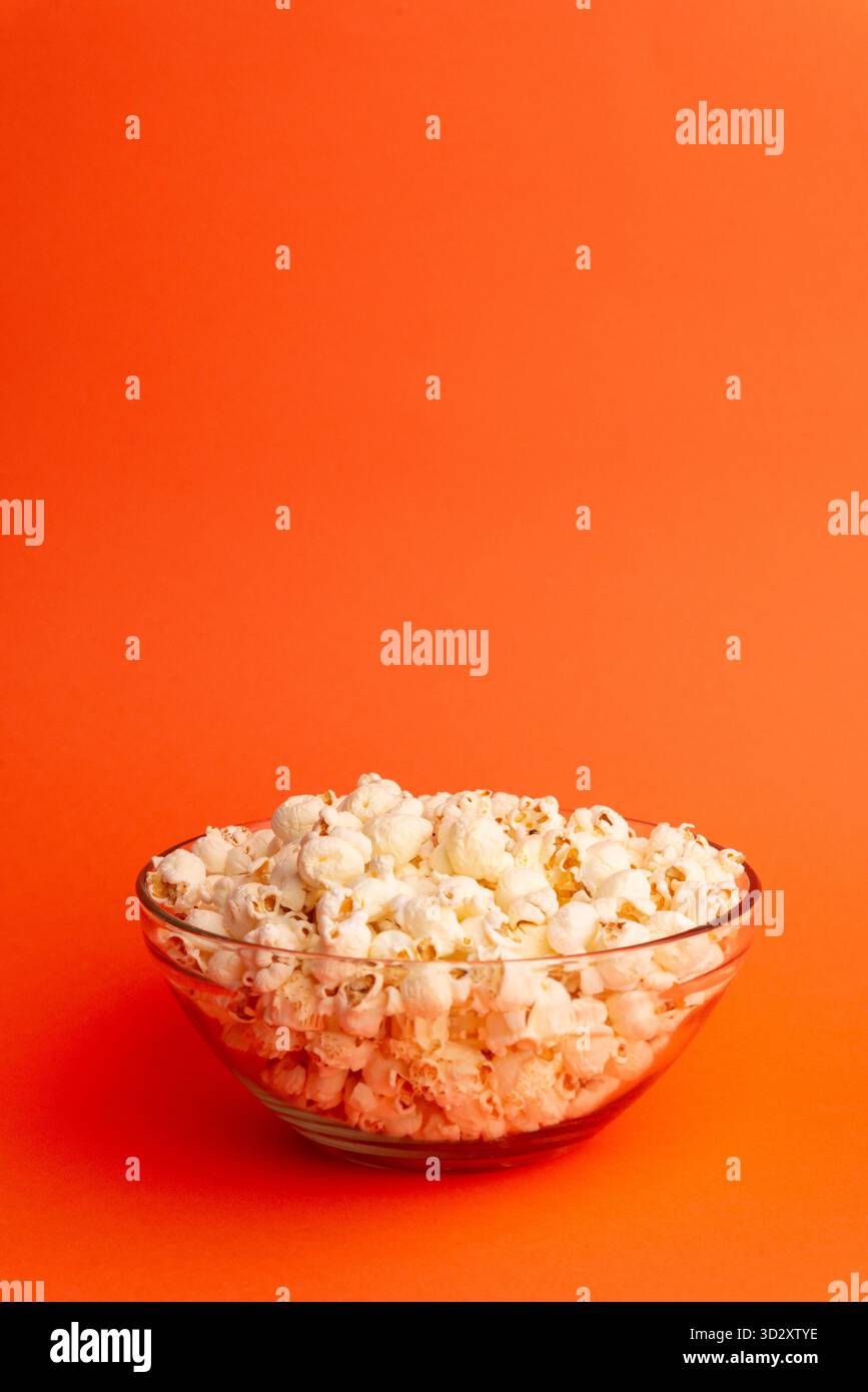 Studio-Foto von Popcorn, das über einer transparenten Schüssel auf orangefarbenem Hintergrund fällt und Bewegung, Lebensmittelkonzept und minimalistisches Kinodesign kombiniert Stockfoto
