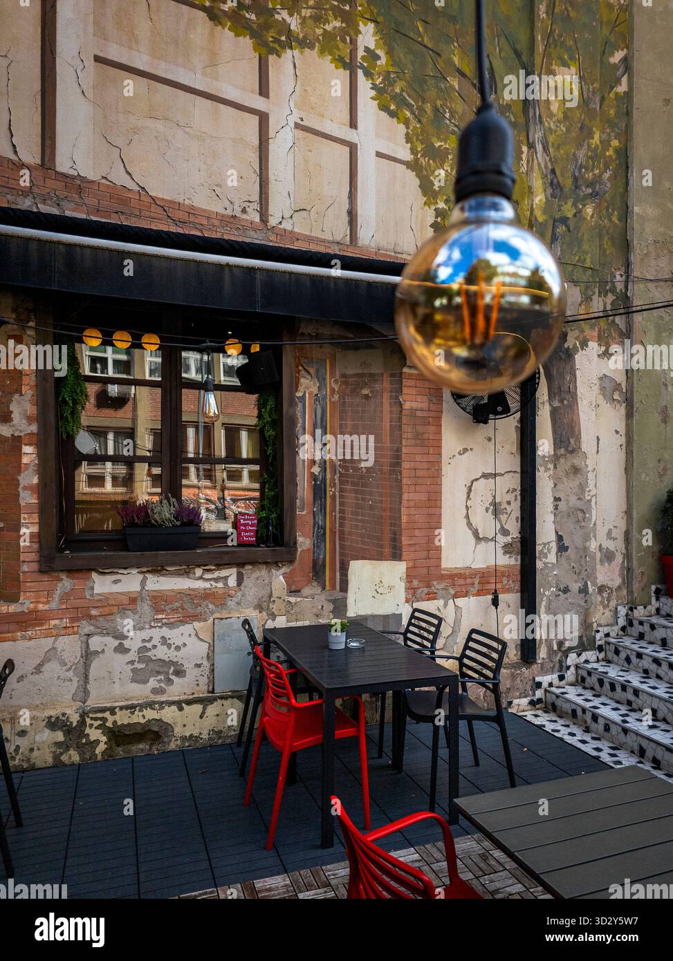 Ein gemütliches Café im Freien in der Skadarska Straße in Belgrad, Serbien, mit roten und schwarzen Stühlen, einer Hängelampe, rustikalen abblätternden Wänden mit bemalten Bäumen, Stockfoto