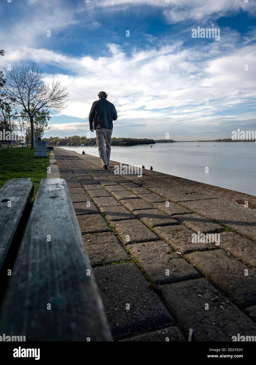 Ein Mann spaziert entlang der Uferpromenade an der Donau in Belgrad, Serbien, unter einem riesigen Himmel, der von weißen Wolken bedeckt ist, mit ruhigem Wasser, weit entfernt Stockfoto