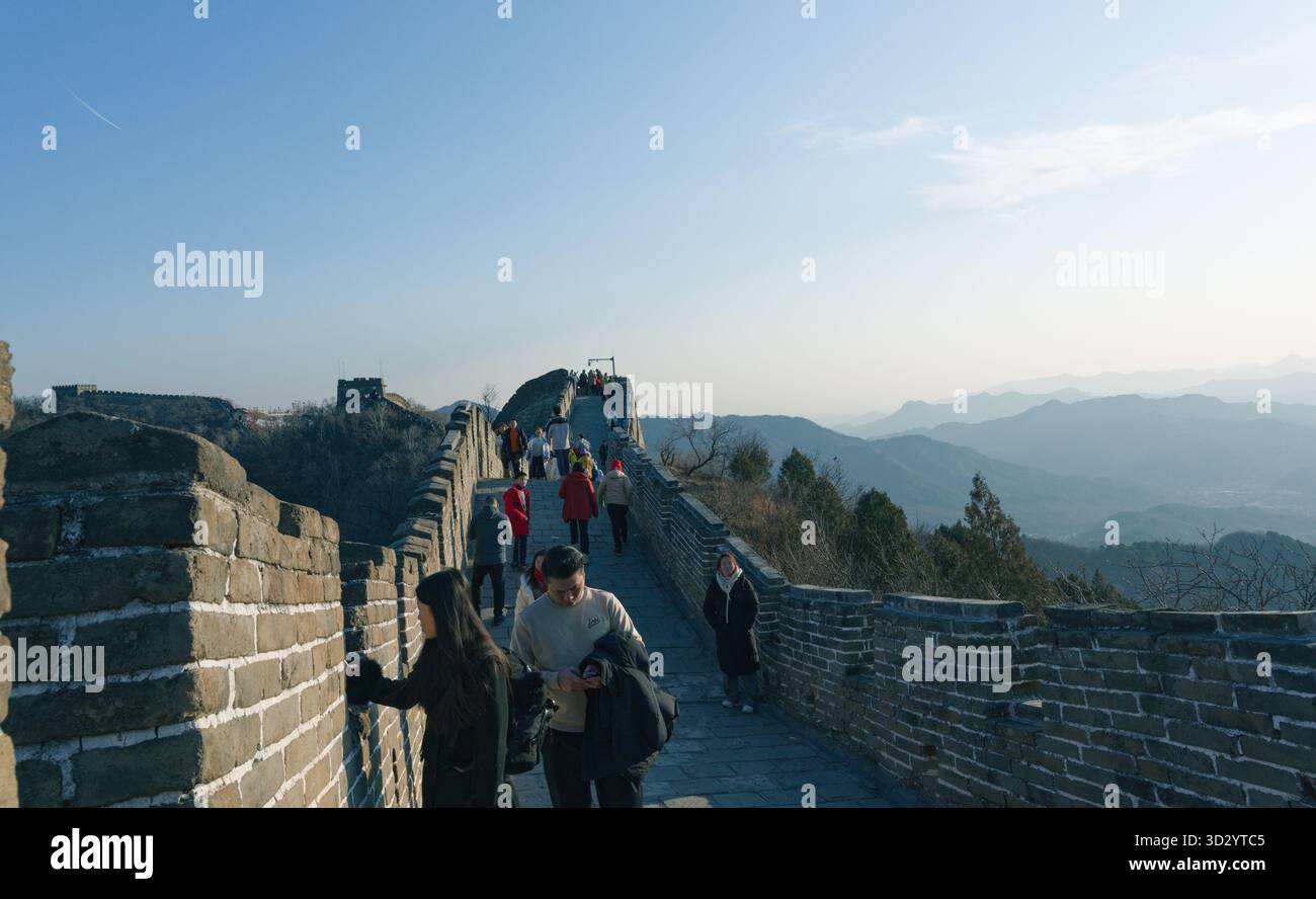 Touristen spazieren entlang eines sonnendurchfluteten Abschnitts der Chinesischen Mauer, einer zerklüfteten Berglandschaft, die sich bis in den trüben Horizont erstreckt Stockfoto
