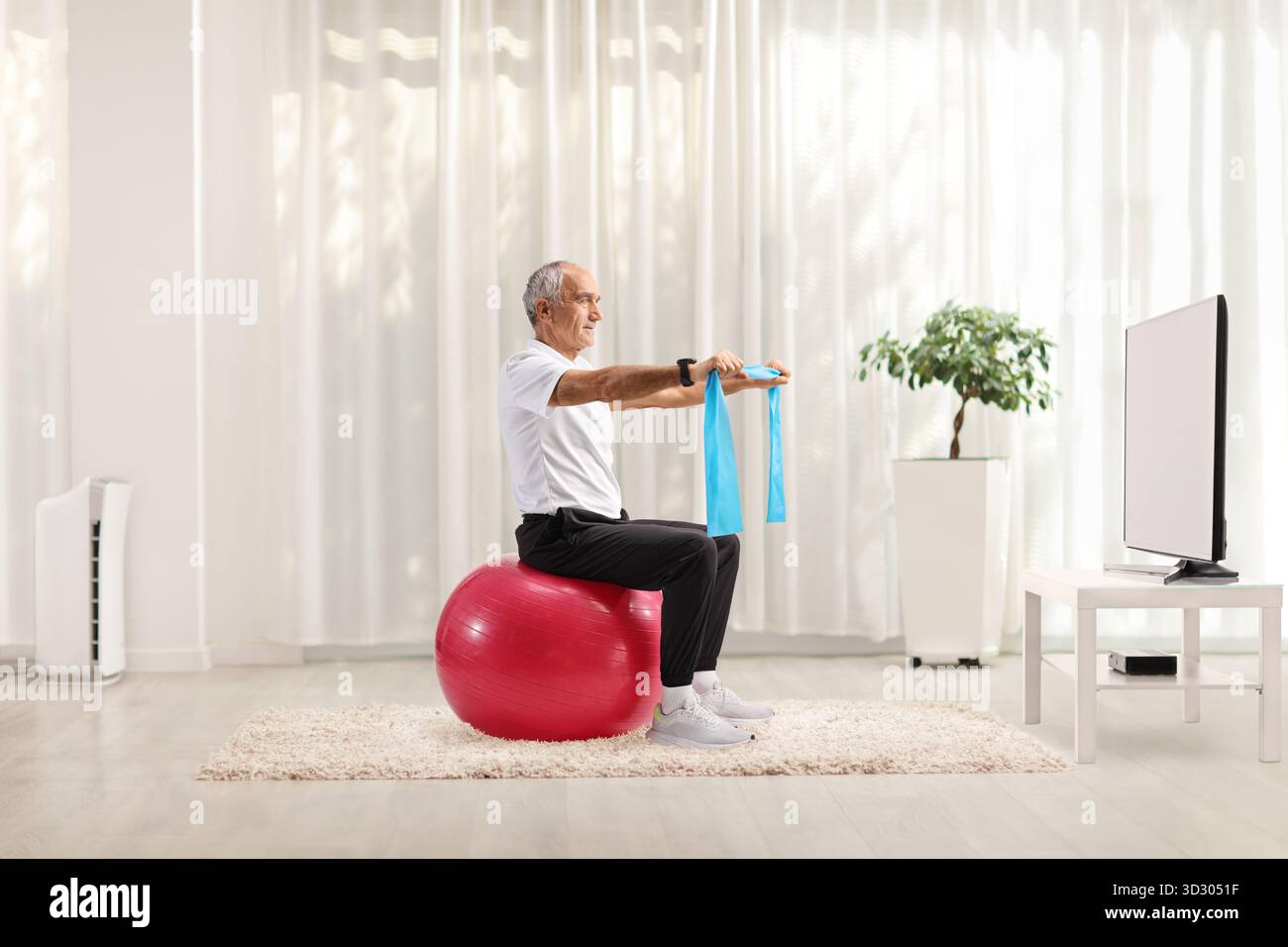 Reifer Mann, der mit einem Widerstandsband trainiert und zu Hause auf einem Fitnessball vor dem fernseher sitzt Stockfoto