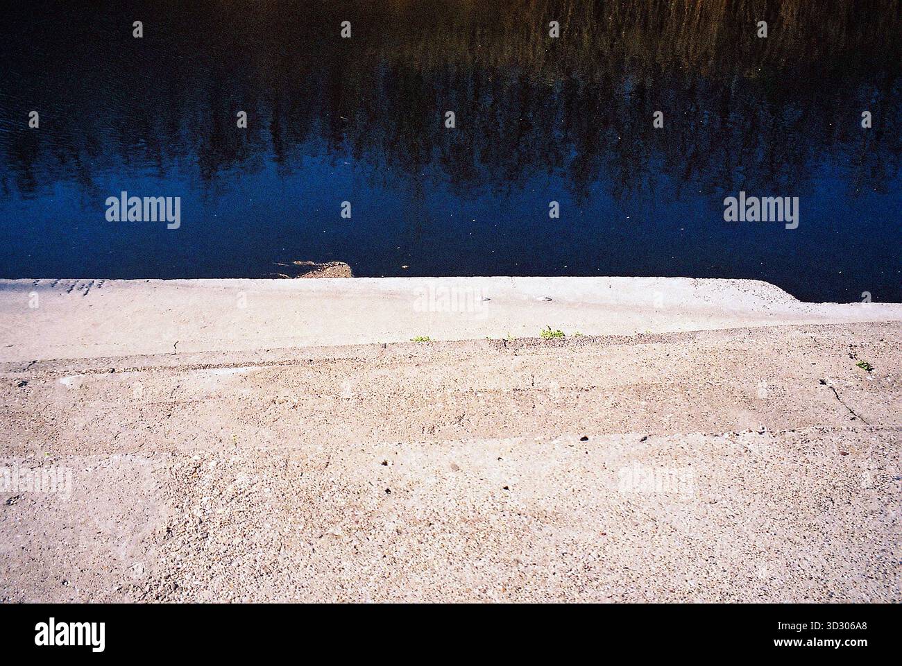 Betonierte Küstenlinie mit ruhigem Wasser, das auf der gegenüberliegenden Seite reflektiert, und einem klaren wolkenlosen Himmel, der in warmen analogen Tönen gehalten wird. Stockfoto