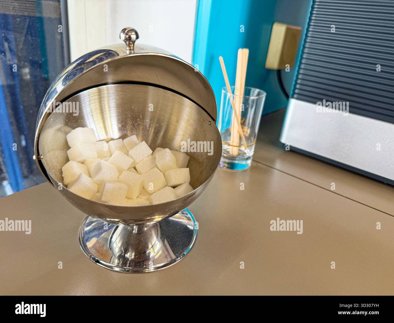 Eine kompakte Metallschale hält weiße Zuckerwürfel unter dem Deckel, die aufgeräumte Portion liegt auf dem Tisch und die einfache Einstellung bietet warme Getränke Stockfoto
