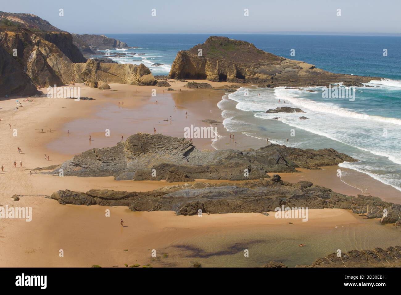 Breiter Sandstrand mit Felsformationen und Schwimmern am Praia da Amália an der portugiesischen Alentejo-Küste, umgeben von zerklüfteten Klippen und Atlantikwellen. Stockfoto