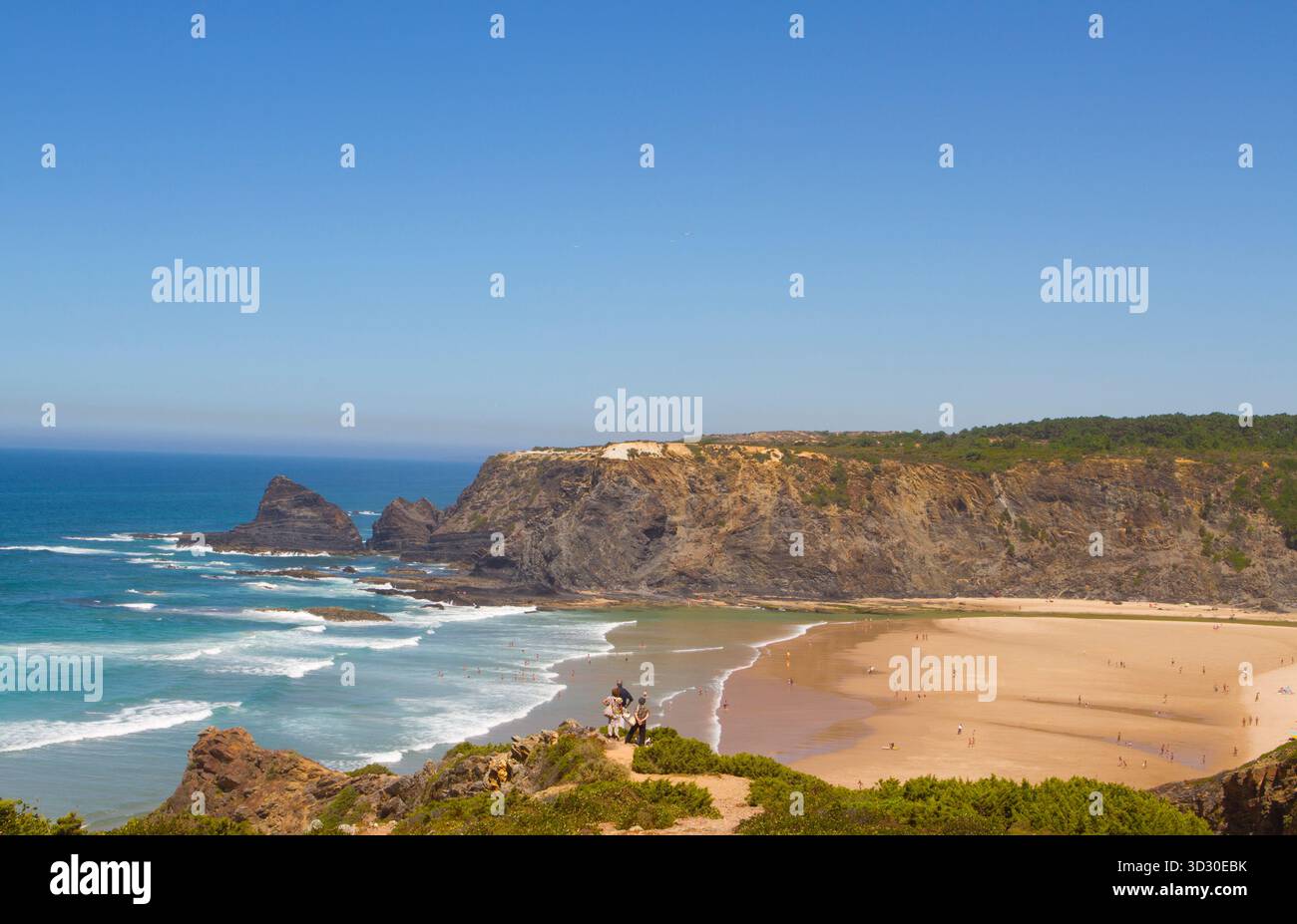 Breiter Sandstrand mit dramatischen Klippen am Praia de Odeceixe an der Küste von Alentejo, Portugal, mit Surfern und Schwimmern im Atlantik. Stockfoto