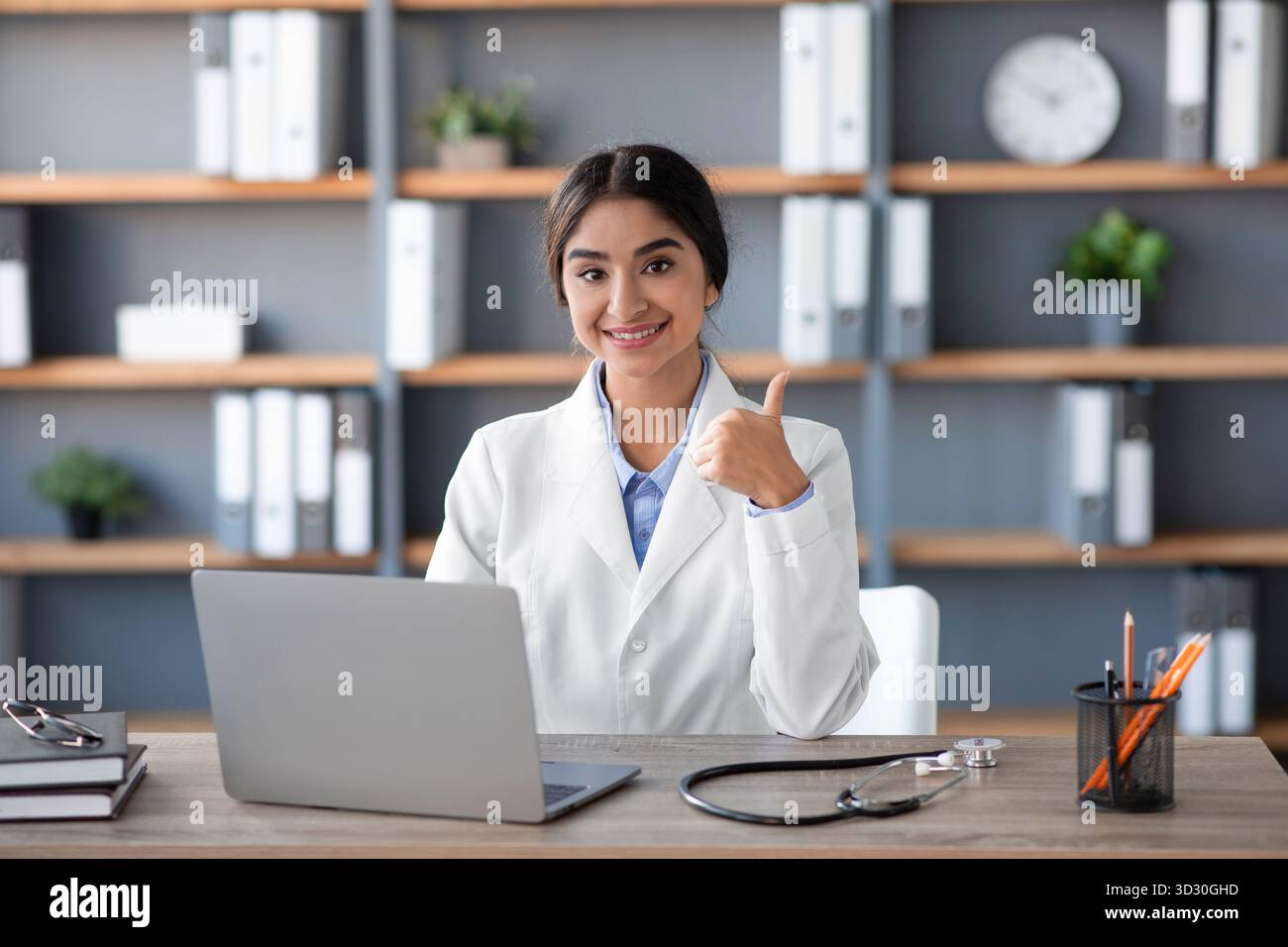 Freundliche indische Ärztin im Klinikbüro gibt Daumen hoch, während sie an Laptop arbeitet Stockfoto