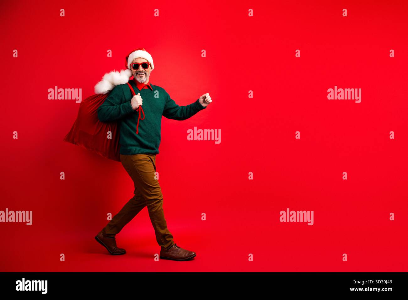 Der lustige Weihnachtsmann trägt eine große rote Tasche mit einem grünen Pullover und einer Sonnenbrille vor einem knallroten Hintergrund Stockfoto